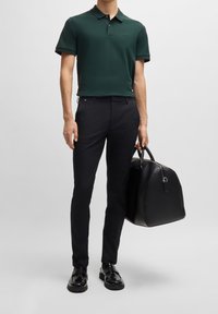 Mørkegrøn polo-shirt med sort kant, sorte slim-fit bukser og polerede sorte sko, holder en struktureret taske i sort læder.