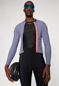 Jersey de ciclismo en púrpura claro con tejido texturizado, con una cremallera frontal con un acento en coral, usado sobre una camiseta base de malla negra. Pantalones cortos de ciclismo negros y un casco.