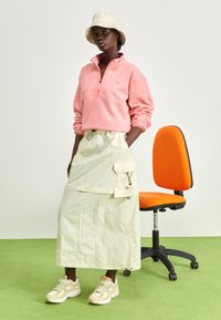 Una mujer se encuentra de pie con las manos en los bolsillos, vestida con un forro polar rosa, una falda cargo beige, un sombrero de cubo y zapatillas, al lado de una silla de oficina naranja sobre un suelo verde.