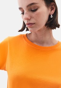 Jasně oranžové tričko s krátkým rukávem vyrobené z měkké tkaniny a kulatým žebrovaným výstřihem. Hladká textura s minimalistickým designem.