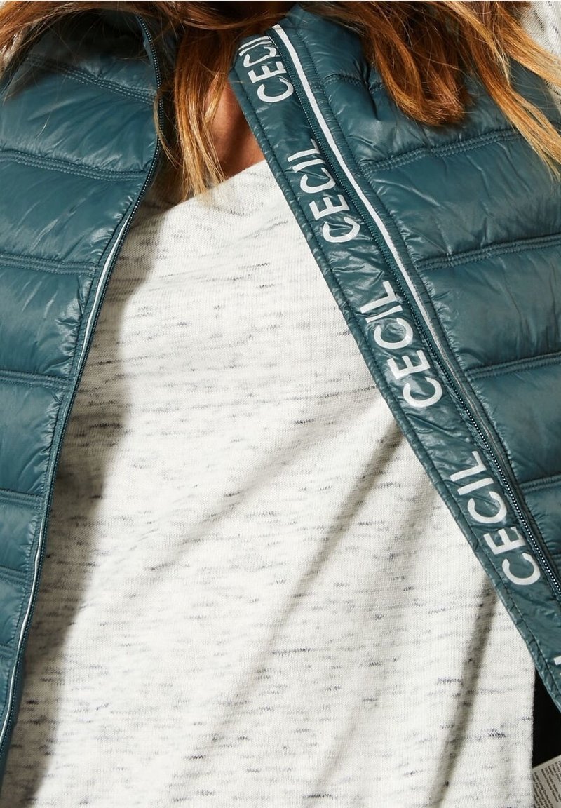 Veste sans manches matelassée teal avec un motif horizontal et un liseré de logo blanc, portée sur une chemise à manches longues gris clair tachetée.