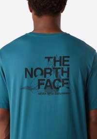 Persona que lleva una camiseta de color aguamarina con el logo negro de "THE NORTH FACE" y un gráfico de montaña, y el texto "NEVER STOP EXPLORING" en la parte de atrás.