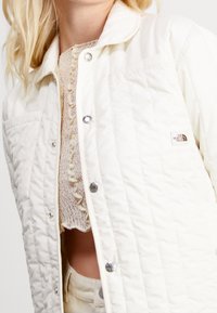 Pehmustettu valkoinen takki, jossa painonapit ja logopäällinen. Kevyt materiaali, jossa teksturoitu pinta ja klassinen kauluksen muotoilu.