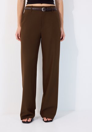 Femme portant un pantalon marron taille haute à jambes larges avec une ceinture noire et des sandales noires à bout carré, debout devant un fond blanc.