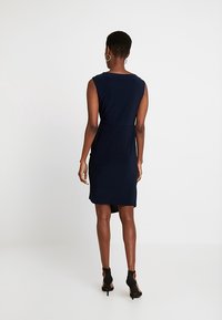 Marinblå ärmlös klänning med en figurnära silhuett, subtil rund halsringning och en mellanhöjd fåll. Bärs med svarta öppna klackar.