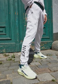 Vita joggerbyxor med en svart "NIKE"-logotyp på sidan, tillsammans med ljusgröna sneakers med mörka accenter och texturerade sulor.