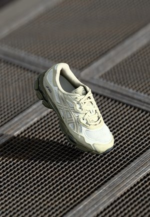 Light beige Asics running shoe floating above metal grate flooring with geometric pattern in soft lighting.