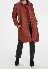 Pikowana długa kurtka w kolorze rdzawego czerwieni, wykonana z błyszczącego materiału syntetycznego, zapinana na zatrzaski, z bocznymi kieszeniami i ściągaczami przy mankietach.