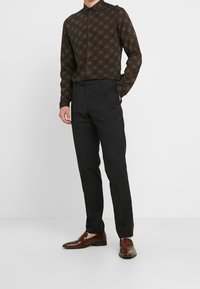 Camicia a maniche lunghe marrone scuro con stampa geometrica, abbinata a pantaloni eleganti neri e scarpe in pelle marroni, in piedi contro uno sfondo bianco.