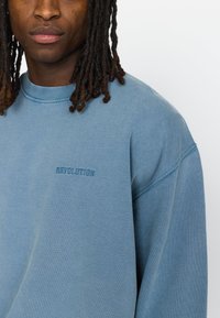 Blauw sweatshirt van zacht materiaal, met een ronde hals en een relaxte pasvorm. "REVOLUTION" is geborduurd in een iets donkerder blauw.