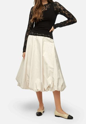 Jupe blanche volumineuse avec un ourlet froncé, associée à un haut en dentelle noire à manches longues. Ballerines couleur tan avec des embouts noirs.