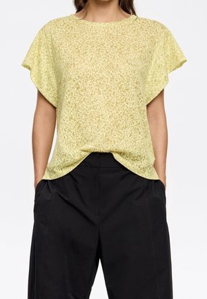 Femme portant un chemisier jaune à manches courtes à motifs, rentré dans un pantalon noir ample taille haute, les mains dans les poches.