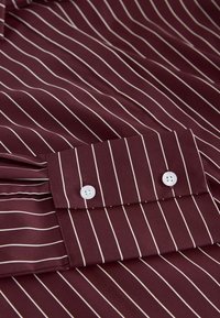 Cămașă bordeaux cu dungi verticale albe, având manșete cu nasturi și o textură de țesătură netedă. Închidere cu două nasturi și cusuturi tonale.