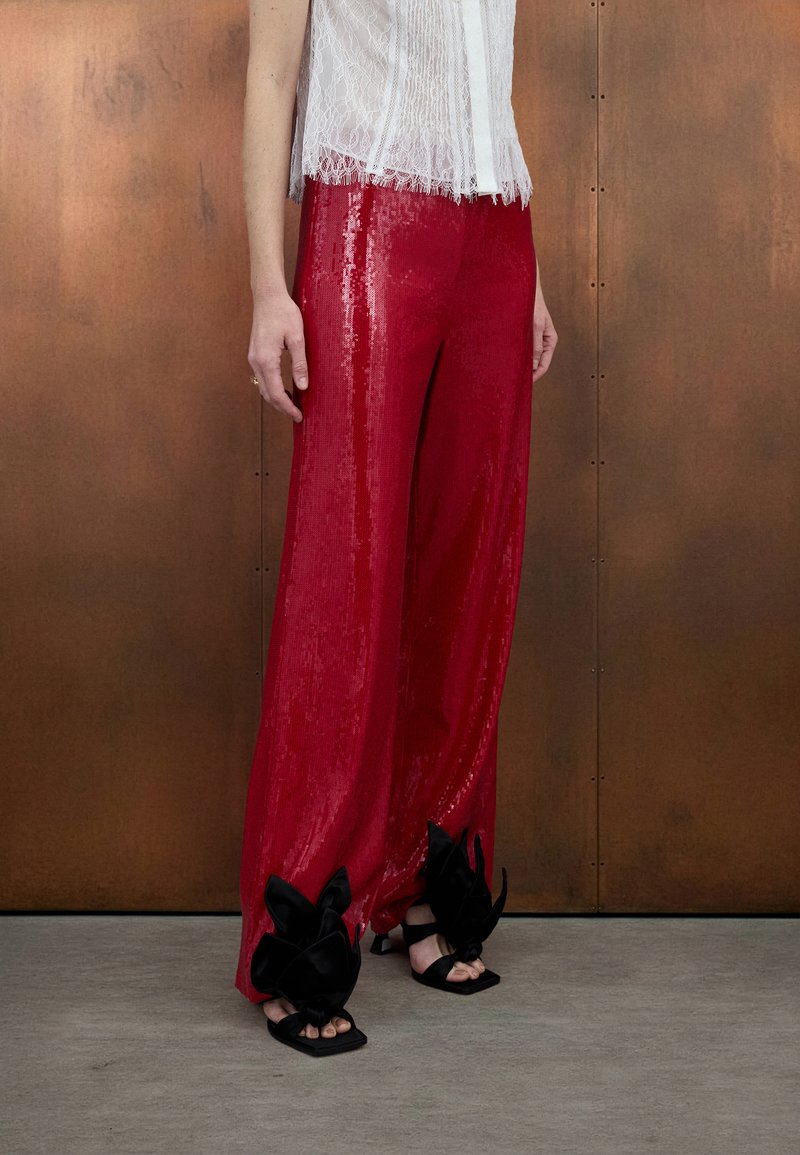 Pantalon en sequins rouges à coupe large, associé à des sandales noires ornées de grands nœuds. Tissu texturé brillant.