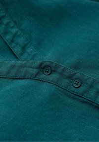 Camisa de lino en color azul verdoso con dos botones oscuros en la tapeta delantera, con una textura suave y un sutil patrón de tejido.