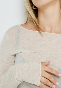 Haut beige clair à manches longues avec un grand col, texture douce et poignets ajustés. Les coutures ajoutent un intérêt visuel supplémentaire.