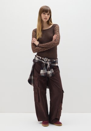 Top marrone in pizzo a maniche lunghe, pantaloni cargo marroni con tasche, camicia a quadri annodata in vita, scarpe bordeaux con suola chiara, sfondo neutro.