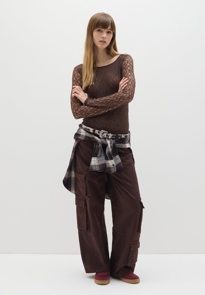 Top marrón de encaje de manga larga, pantalones cargo marrones con bolsillos, camisa de cuadros atada a la cintura, zapatos burdeos con suela clara, fondo neutro.