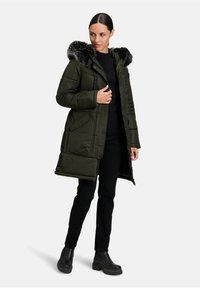 Zielona kurtka puchowa z kapturem wykończonym sztucznym futrem, zamkiem błyskawicznym z przodu i dwoma bocznymi kieszeniami. Nosi się ją na czarną bluzkę i spodnie. Masywne czarne botki.