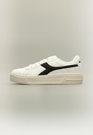 Baskets basses blanches avec semelle texturée et motif noir en forme de flèche sur le côté, présentées sur un fond neutre.