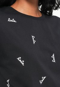 Svart T-skjorte med korte ermer og rund hals, med tilfeldig plasserte hvite broderte "lala"-tekster i kursiv font over stoffet.