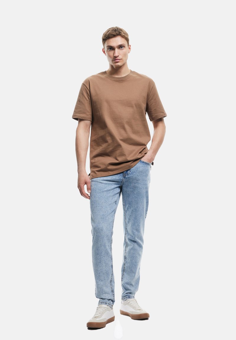 Camiseta de algodón marrón de manga corta, combinada con vaqueros de mezclilla azul claro y zapatillas blancas con detalles marrones. Textura suave.