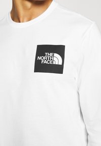 Weißes Langarm-T-Shirt mit einem schwarzen quadratischen Logo auf der linken Brust, das den Text "THE NORTH FACE" und ein stilisiertes Bogen-Design zeigt.