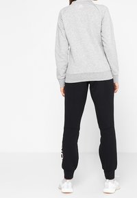 Felpa grigia con colletto alto, polsini e orlo a coste. Pantaloni jogger neri con dettaglio del logo sulla gamba. Scarpe da ginnastica completano il look.
