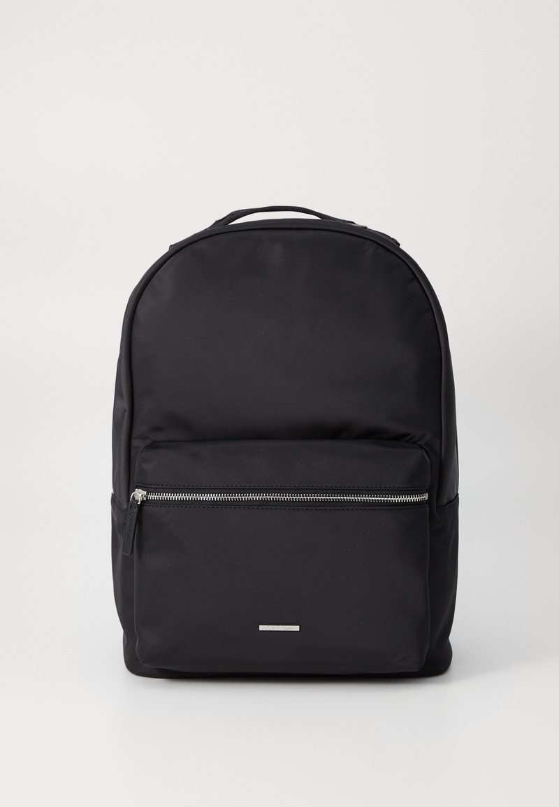 Mochila negra hecha de tela duradera, con una superficie suave, diseño redondeado, un bolsillo frontal con cremallera y un acento plateado minimalista.