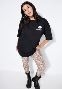 Camiseta negra oversize con logo gráfico, combinada con leggings de estampado de leopardo beige y botas chunky negras. El modelo posa contra un fondo blanco.