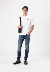 T-shirt blanc avec logo rouge Diesel, jean bleu slim-fit et chaussures en cuir noires. Sac bandoulière noir avec sangle réglable.