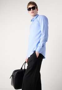 Camisa de botones azul claro con mangas largas, combinada con pantalones negros. El modelo sostiene un bolso de cuero negro, vistiendo gafas de sol oscuras.