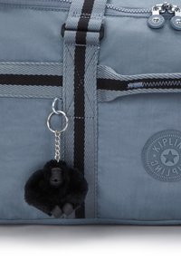 Sininen kankainen laukku mustaraitaisilla hengittimillä, vetoketjulla, pyöreällä Kipling-logolla ja pienellä mustalla karvaisella apinapatsalla, joka on kiinnitetty olkaremmin silmukkaan.