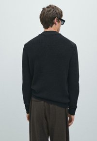 Maglione nero a maglia con finitura testurizzata, colletto rotondo e maniche lunghe, abbinato a pantaloni marroni. Vista posteriore, design minimale.