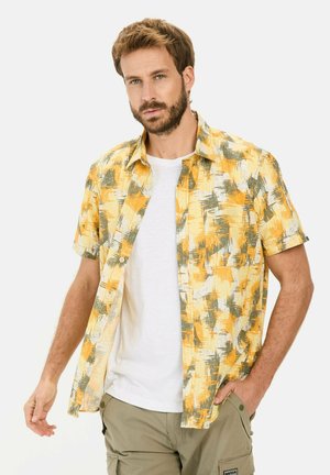 Kurzärmliges, knöpfbares Hemd in einem abstrakten Muster in Gelb, Grün und Weiß; leichtes Gewebe mit lockerer Passform, über einem weißen T-Shirt getragen.