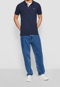 Mörkblå pikétröja med tvåknappsslutning, med röd och vit detaljer. Matchas med löst sittande blå jeans och vita sneakers.