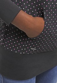Sudadera negra con un patrón de puntos en rosa y blanco, con un bolsillo frontal y un dobladillo inferior acanalado. Textura de tela suave y ajuste casual.