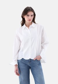 Weißes Hemd mit Kragen und langen Ärmeln, das einen leicht geschwungenen Saum und subtile vertikale Nähte aufweist. Getragen mit blauen Jeans.
