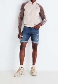 Hellbeige und braune Adidas-Jacke mit Reißverschluss und drei Streifen an den Ärmeln. Blaue Denim-Shorts mit Distressed-Details. Beigefarbene Schuhe mit Ausschnitten.