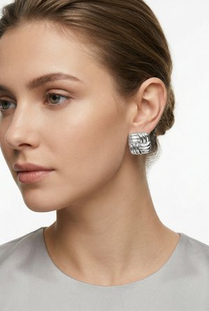 Vaaleaihoinen ja vihreäsilmäinen nainen käyttää neliönmuotoisia hopeisia kudottuja korvakoruja ja vaaleanharmaata yläosaa, katsoo vasemmalle yksinkertaista valkoista taustaa vasten.