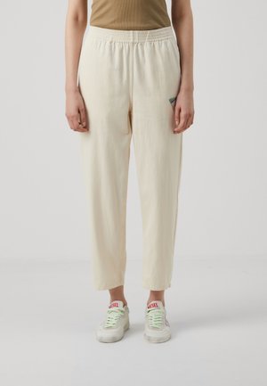 Personne portant un pantalon ample couleur crème avec une taille élastique et des baskets blanches Diesel, debout sur un fond clair uni.
