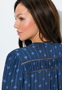 Marineblauwe blouse met hartjespatroon, voorzien van een geplooide achterkant en transparante banddetails. Gladde textuur, losse pasvorm.