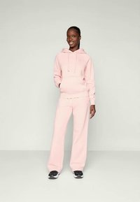 Felpa rosa chiaro con tasca frontale e coulisse, abbinata a pantaloni della tuta rosa coordinati. Il tessuto sembra morbido e testurizzato. Completato con sneaker nere.