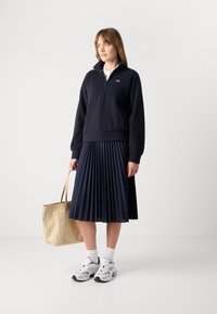 Navy zip-up sweatshirt with long sleeves, paired with a pleated navy skirt. Model carries a beige tote bag and wears white sneakers with black accents.