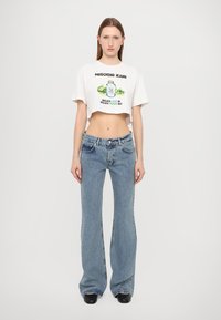 Hvit avkortet t-skjorte med et fargerikt grafisk trykk og tekst, kombinert med lyseblå buede jeans og svarte sko.