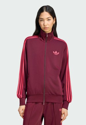 Frau mit langen dunklen Haaren, die eine bordeauxrote Trainingsjacke mit Reißverschluss und passende Hosen mit pinken Streifen und Logos trägt, vor einem schlichten Hintergrund stehend.