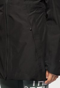 Veste noire zippée avec poches avant portée par une personne portant des leggings foncés avec un texte blanc sur la cuisse.