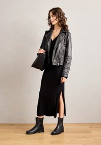 Žena v černé kožené bundě, černých midi šatech s postranním rozparkem, černých kotníkových botách a držící černou kabelku, stojící na dřevěné podlaze.