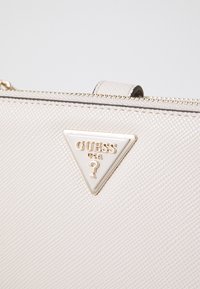 Borsa a tracolla in ecopelle testurizzata di colore crema, con il logo triangolare Guess in oro. Chiusura con zip, design snello e una leggera lucentezza.