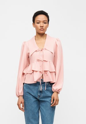 Vrouw draagt een lichtroze blouse met lange mouwen, versierd met ruches en strikken, gecombineerd met een blauwe jeans met hoge taille, staand tegen een effen achtergrond.